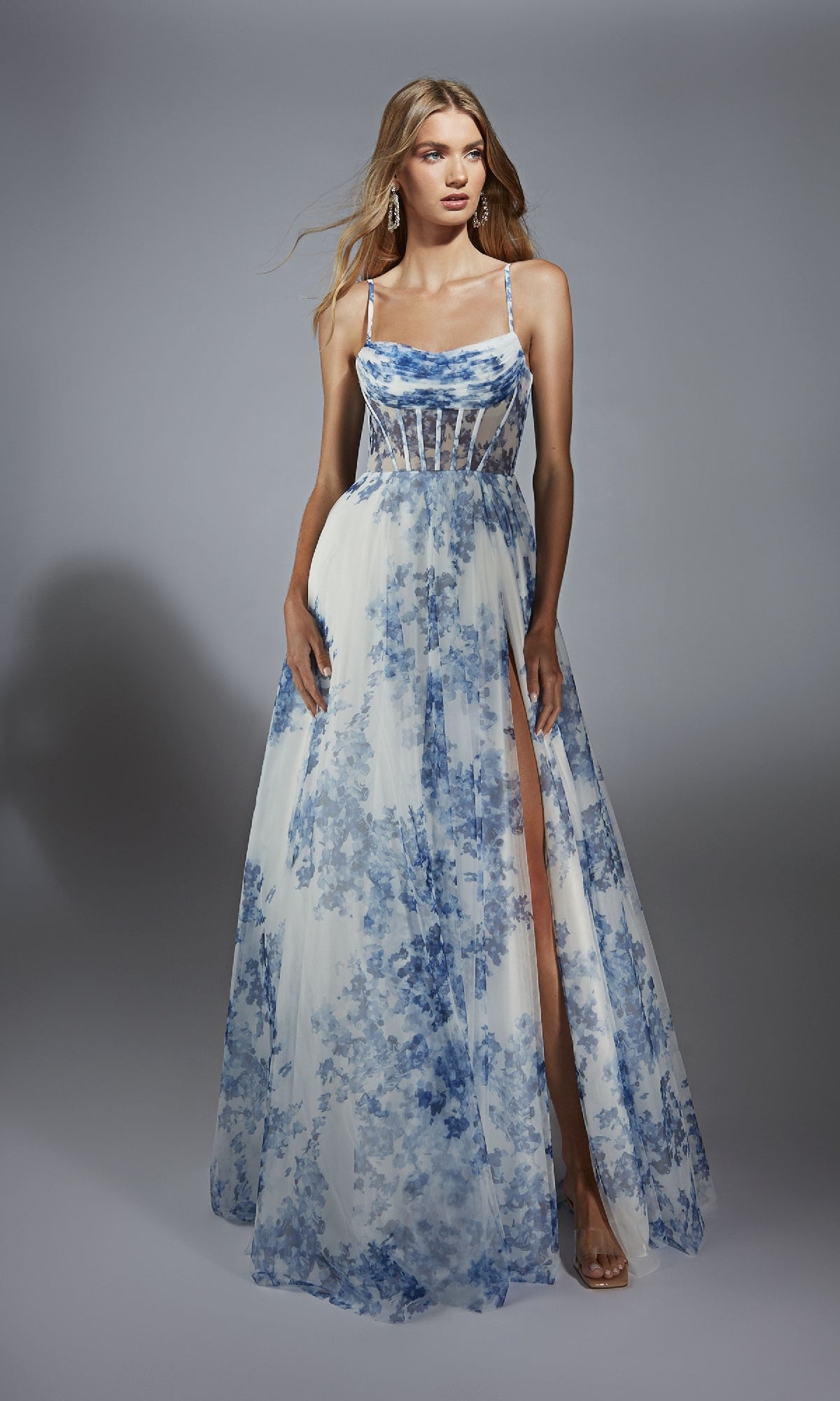 A woman models the Alyce 61752 long prom dress with blue and white floral print, corset bodice, spaghetti straps, and a high leg slit, set against a plain gray background.